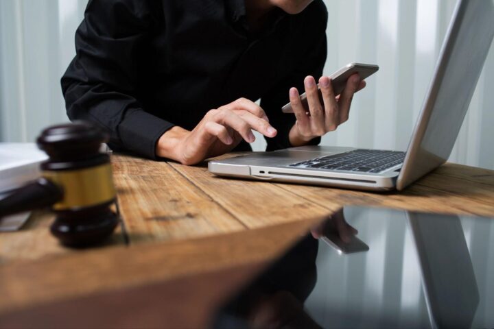 A lawyer or accountant in a black button-down shirt works with a smartphone in one hand, bending over a laptop computer at a desk that also holds a gavel; concept image for tracing hidden assets in a divorce.