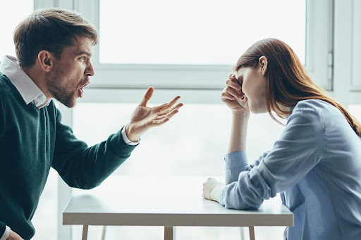 Man yelling across a bare table at a woman who rests her head in one hand; concept for seeking mediation in high conflict divorce.