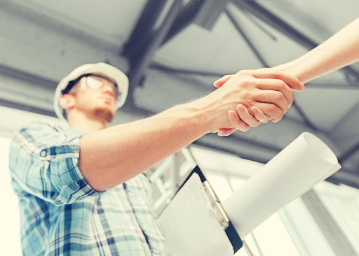 Building contractor holding building plans and clipboard, shaking hands with homeowner; concept for verifying credentials to prevent contractor fraud.