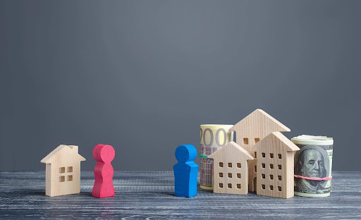 Wooden figures to indicate former spouses, with the female figure standing beside a small wooden house and the male figure standing beside three wooden buildings and two rolls of cash. Hidden assets in high net worth divorces can result in an unfair division of property.