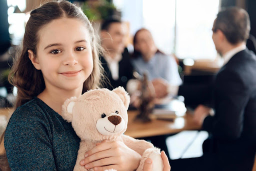 A child hugging a teddy bear while her parents discuss family mediation for blended families with an attorney.