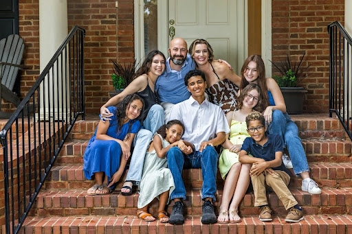 Large blended family with two parents and seven children sitting on the front steps of a brick house; stepchild adoption can legally codify your commitment to the children your spouse had before your marriage.